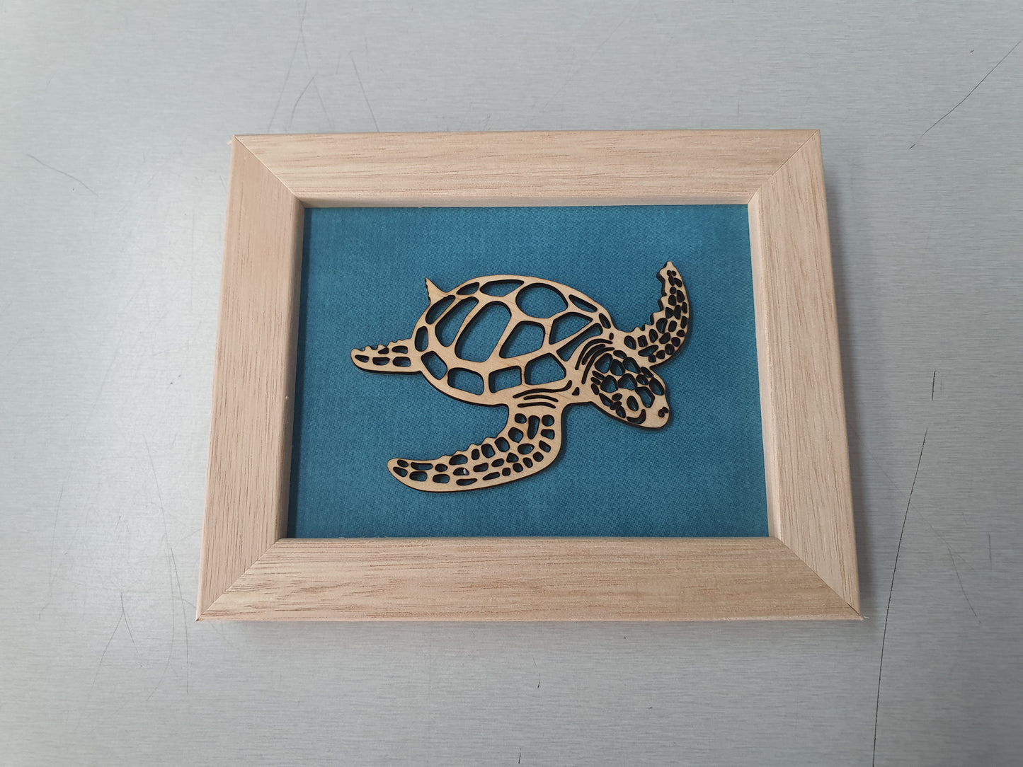 Wooden turtle cutout on a teal background in a light wood frame.